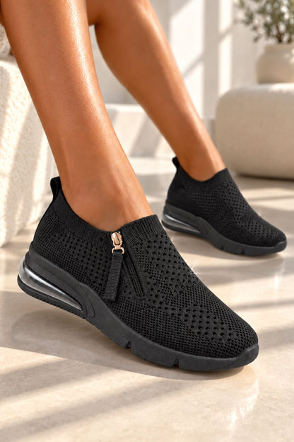 A person wearing black slip-on knit sneakers with a side zipper and tassel detail, shown in a bright indoor setting with sunlight and a light stone floor.