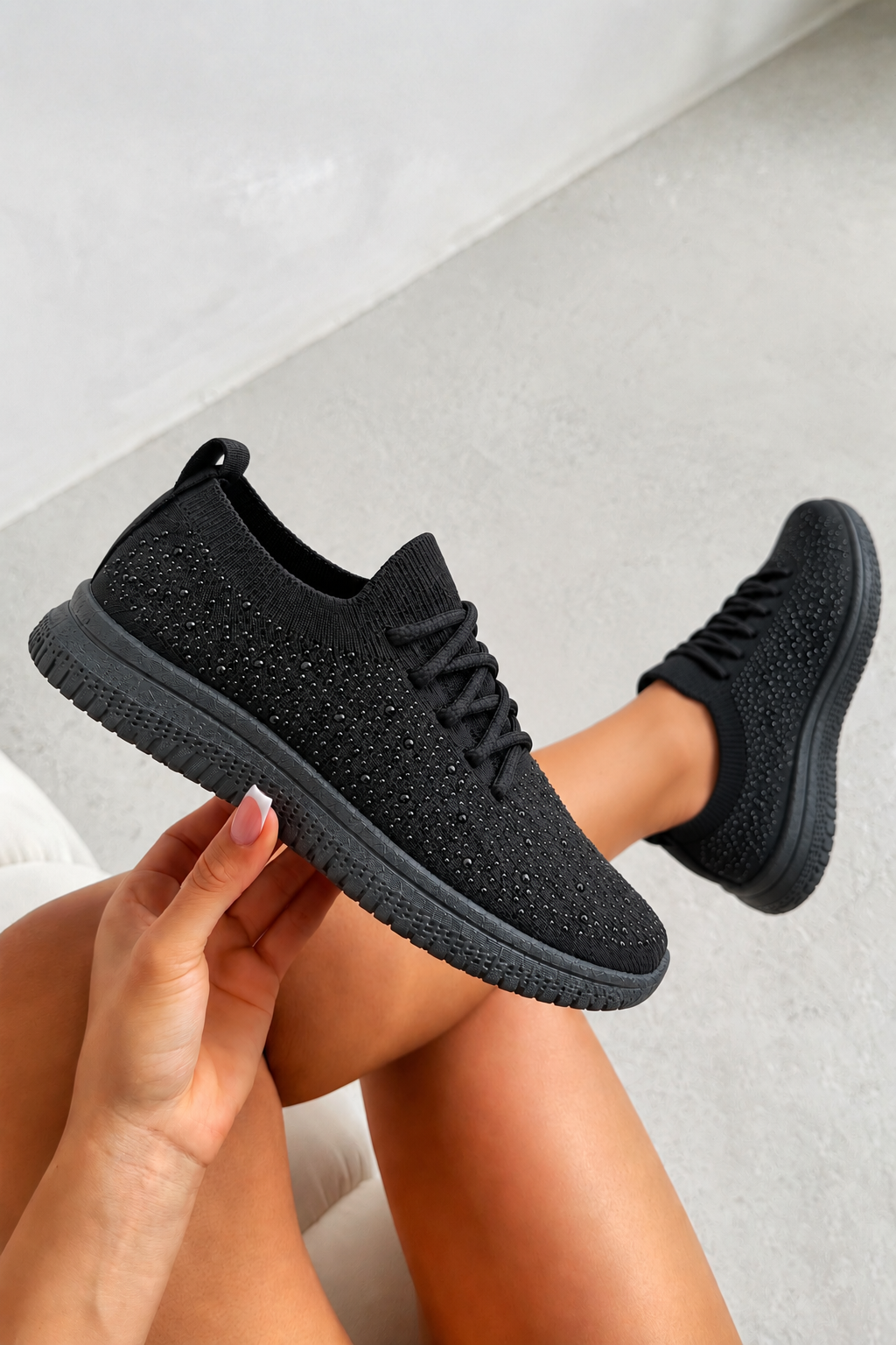 A pair of black knit slip-on sneakers with textured soles, shown being worn with one shoe held up by a hand against a light gray concrete background.