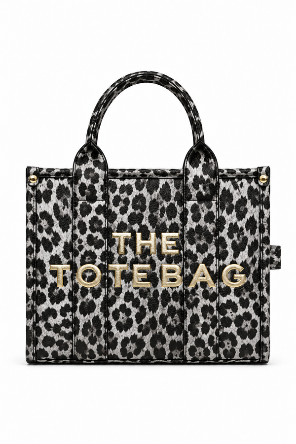 Leopard-print tote bag with a black and grey animal print, two matching top handles, structured rectangular shape and bold gold-tone. "THE TOTE BAG" lettering on the front. Set against a white background. 