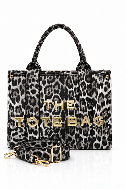 Grey and black Leopard-print tote bag with animal print, two matching top handles, structured rectangular shape and bold gold-tone. "THE TOTE BAG" lettering on the front. Handle of bag pictured in front of bag. Set against a white background. 