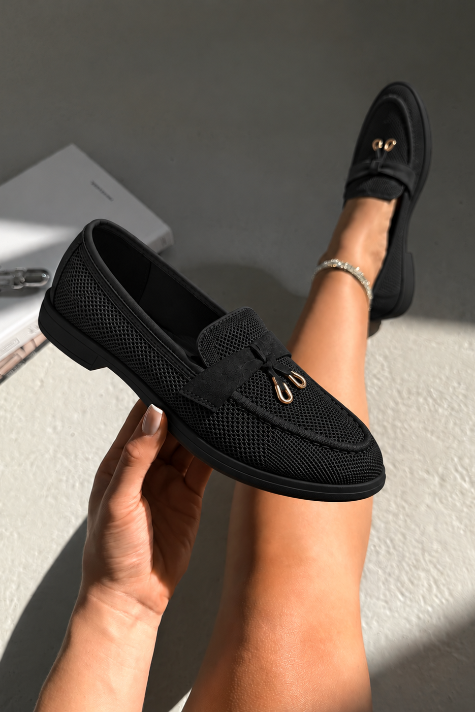 A person holds a black mesh loafer in one hand while wearing the same loafer on their foot, set against a neutral gray background with a book and jewelry visible.