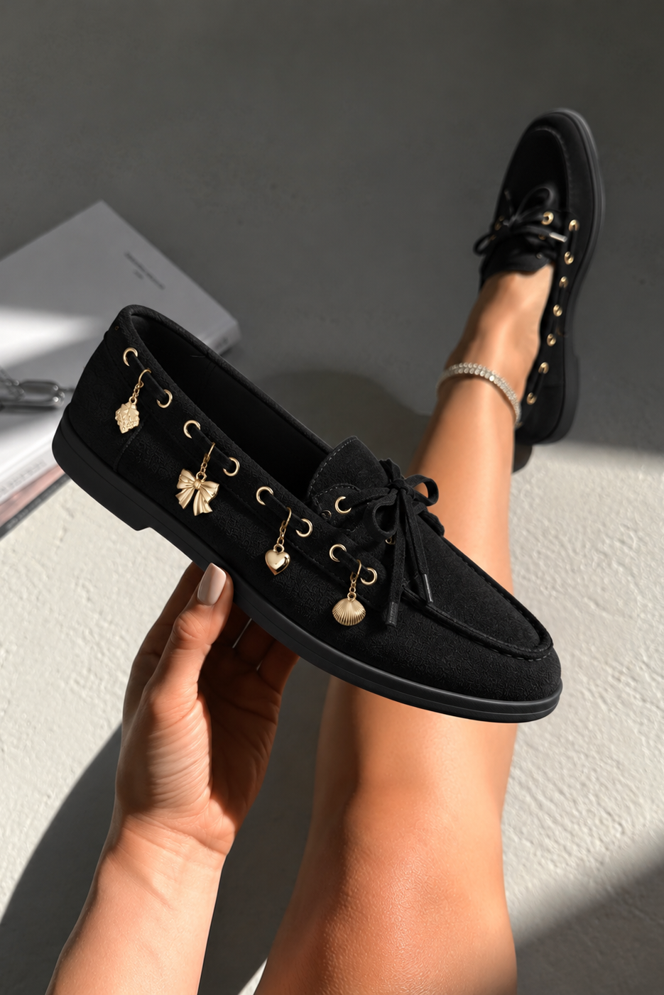 Black suede boat shoes with gold-tone decorative charms, shown being held by a hand and worn on a foot, set against a textured gray wall with natural sunlight and shadows.