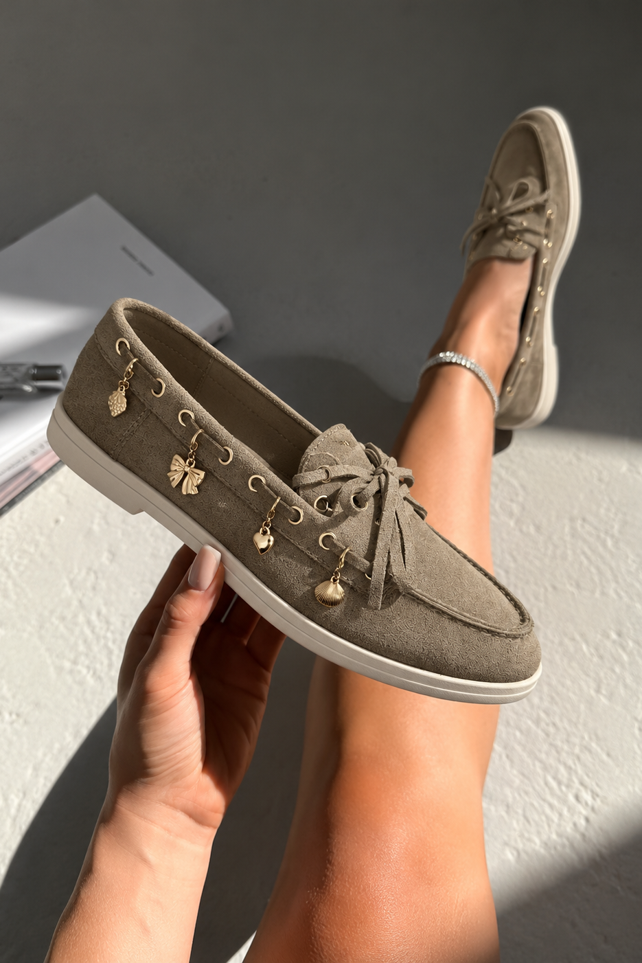 A person holds one taupe-brown suede boat shoe in their hand, with the other shoe worn on their foot. The shoes feature gold-tone decorative charms and gold eyelets, white soles, and matching brown laces.