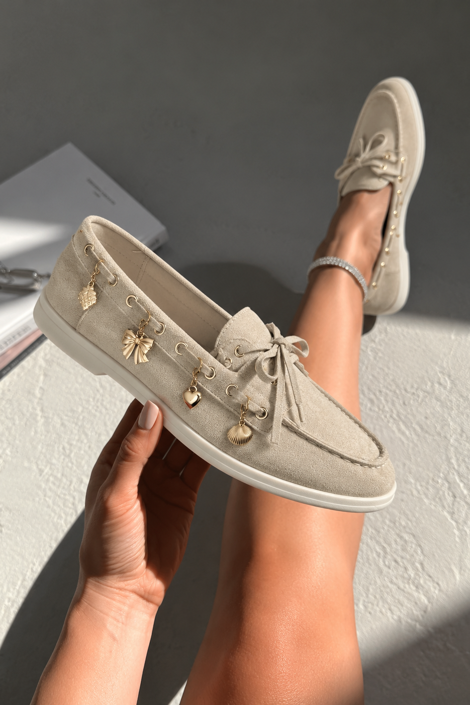 A person holds up a beige suede moccasin-style loafer with dangling gold charms, while wearing the matching shoe on their foot. The scene is set on a textured gray floor with a book in the background.