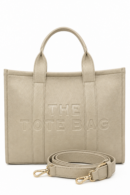 Large Beige faux leather structured tote bag with two rounded top handles, embossed. "THE TOTE BAG" lettering on the front, vertical stitched panels. Pictured on a white background.  With beige-tone strap pictured in front of the bag.