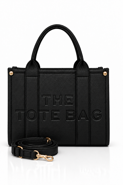 Black faux leather tote bag with two rounded top handles, embossed "THE TOTE BAG" lettering on the front, contrast stitching, gold tone hardware and a detachable adjustable black shoulder strap.