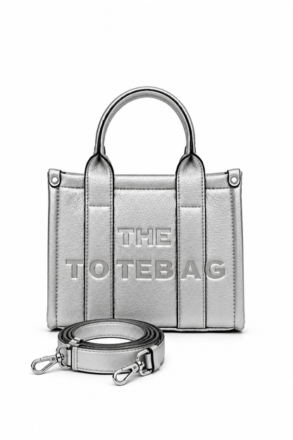 Beige mini faux leather structured tote bag with two rounded top handles, vertical stitched panels, matching tonal stitching and subtle embossed "THE TOTE BAG" lettering on the front. Silver shoulder strap pictured in front of bag. 