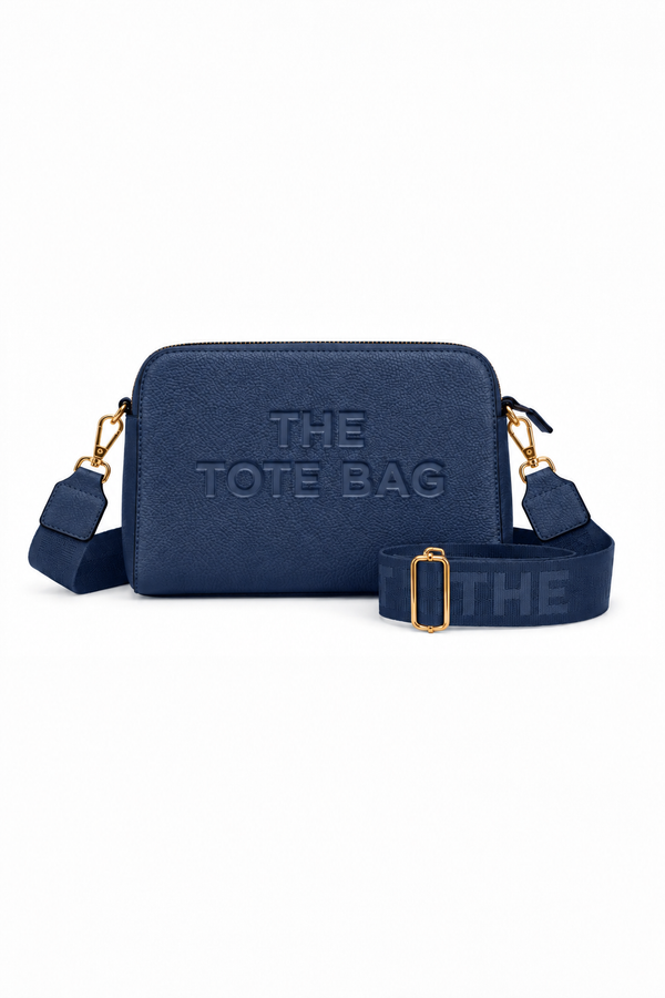 Navy Blue cross body bag with a rectangular shape, rounded corners, embossed, "THE TOTE BAG" lettering on the front, gold-tone clips and a wide adjustable matching shoulder strap with a gold tone buckle. 