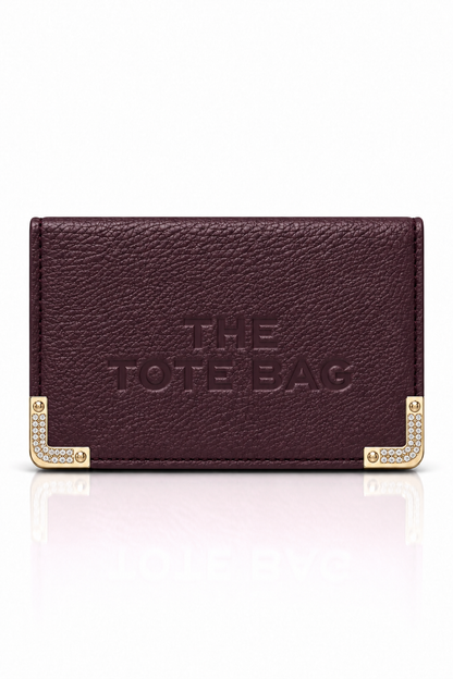 Plum textured tote purse with embossed "The Tote Bag" lettering on the front and cream and gold-tone decorative metal corner protectors with stud detailing. Set against a plain white background.