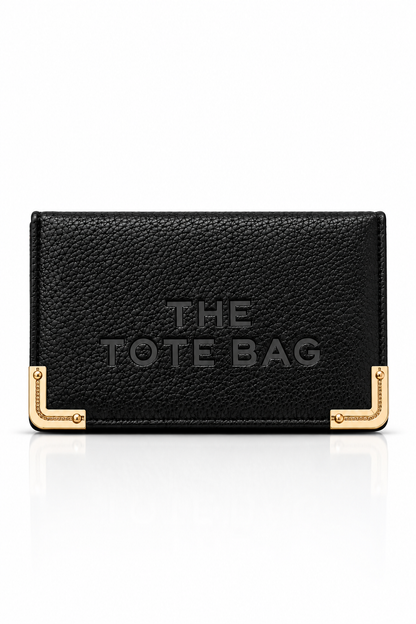 Black textured faux leather purse featuring THE TOTE BAG lettering on the front, with contrasting gold tone metal corner protectors and decorative detailing. Set against a plain white background.