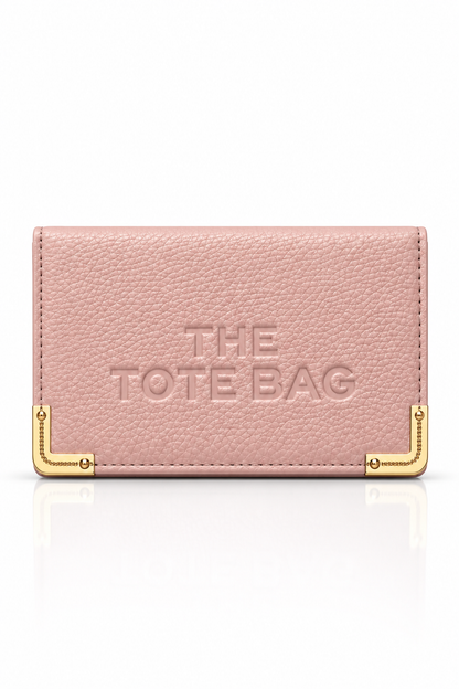 Light pink textured tote bag purse with embossed "THE TOTE BAG" lettering on the front, featuring gold tone metal corner protectors with decorative stud detailing. 