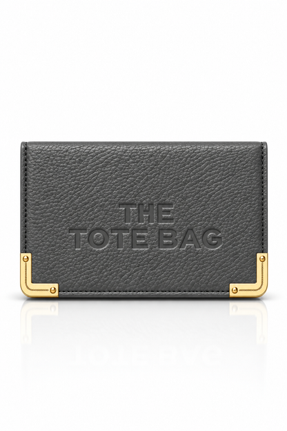 Charcoal grey textured tote purse with embossed "The Tote Bag" lettering on the front and gold tone metal corner protectors with rivet detailing. 