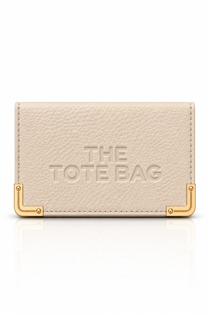 Nude faux leather Tote purse with embossed The Tote Bag lettering on front, textured finish, and gold-tone metal corner protectors with visible rivet detailing.Set against a plain white background.