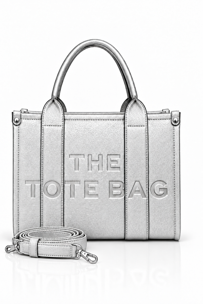 Metallic Silver structured tote bag with two rounded top handles, embossed. "THE TOTE BAG" lettering on the front, vertical stitched panels, contrast edging and silver- tone stud details. With silver-tone strap pictured in front of the bag.