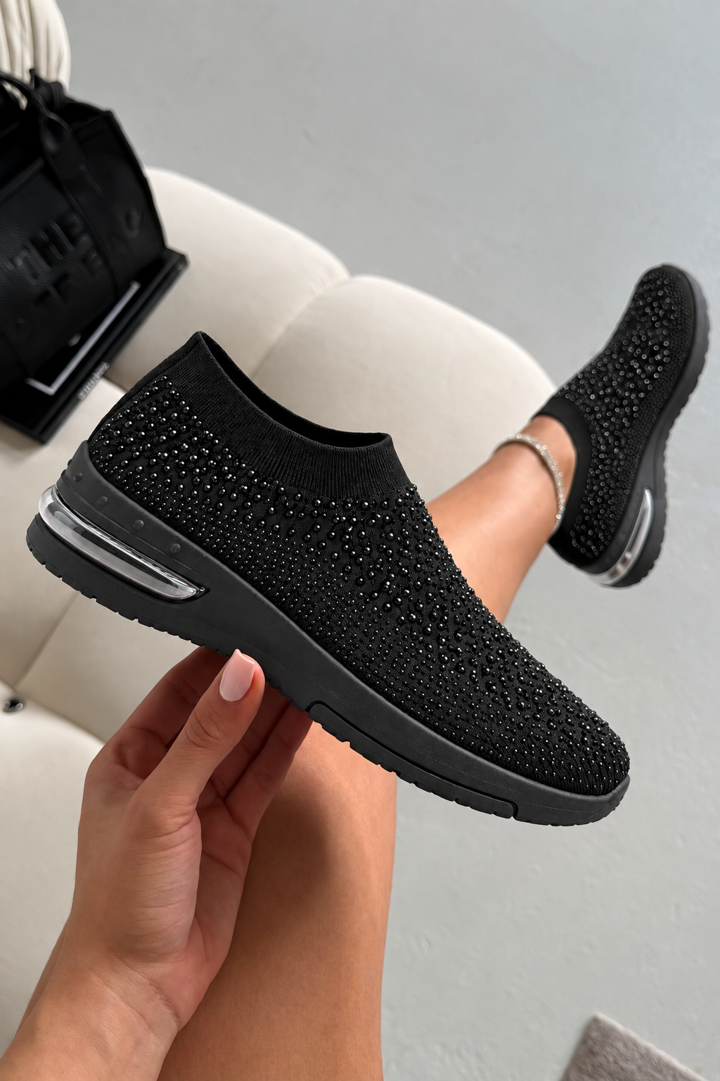 A person holds up a black slip-on sneaker with rhinestone-like embellishments, while wearing the same shoe on one foot. The shoe has a knit upper, a chunky black sole, and a visible air unit on the side, photographed in an indoor setting with a light-colored chair and bag in the background.