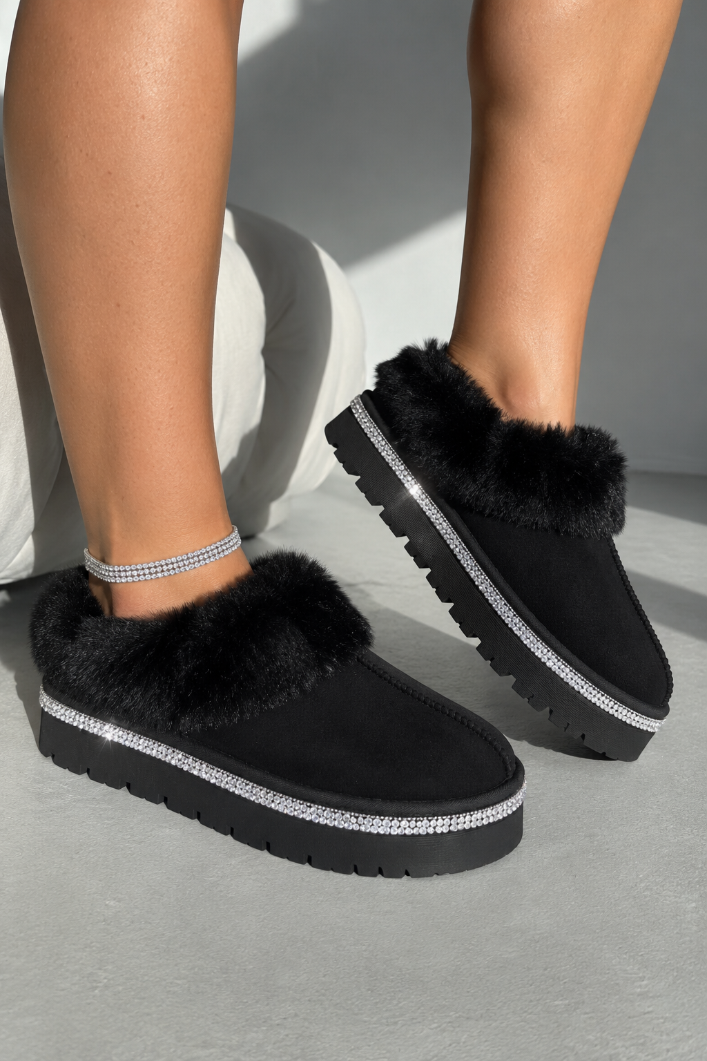A person wearing black slip-on platform slippers with fluffy trim and a silver rhinestone-embellished band, photographed against a light gray studio background.