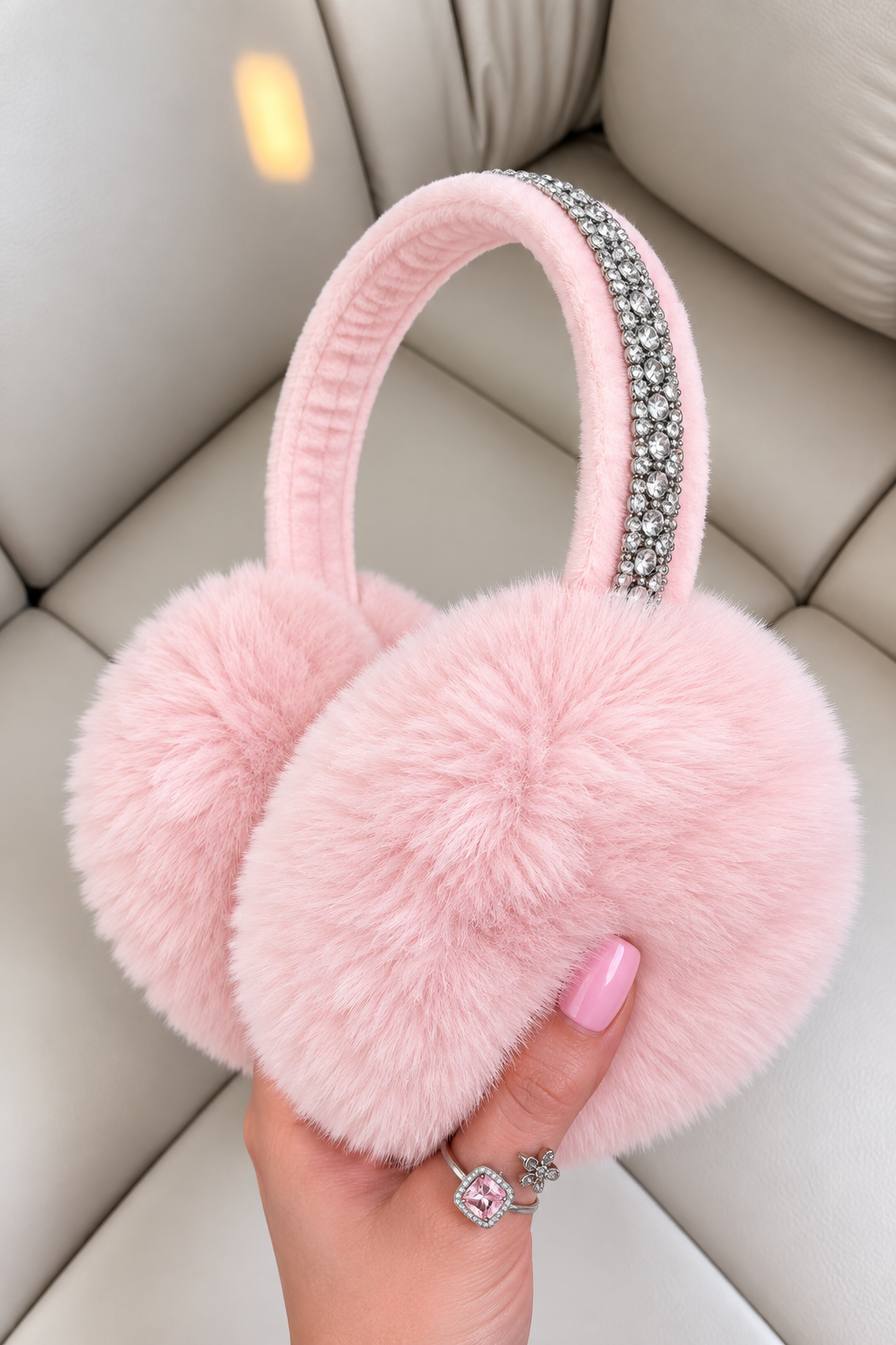 Pink faux fur earmuffs with a rhinestone-embellished headband, held by a hand with pink nail polish against a beige leather background.