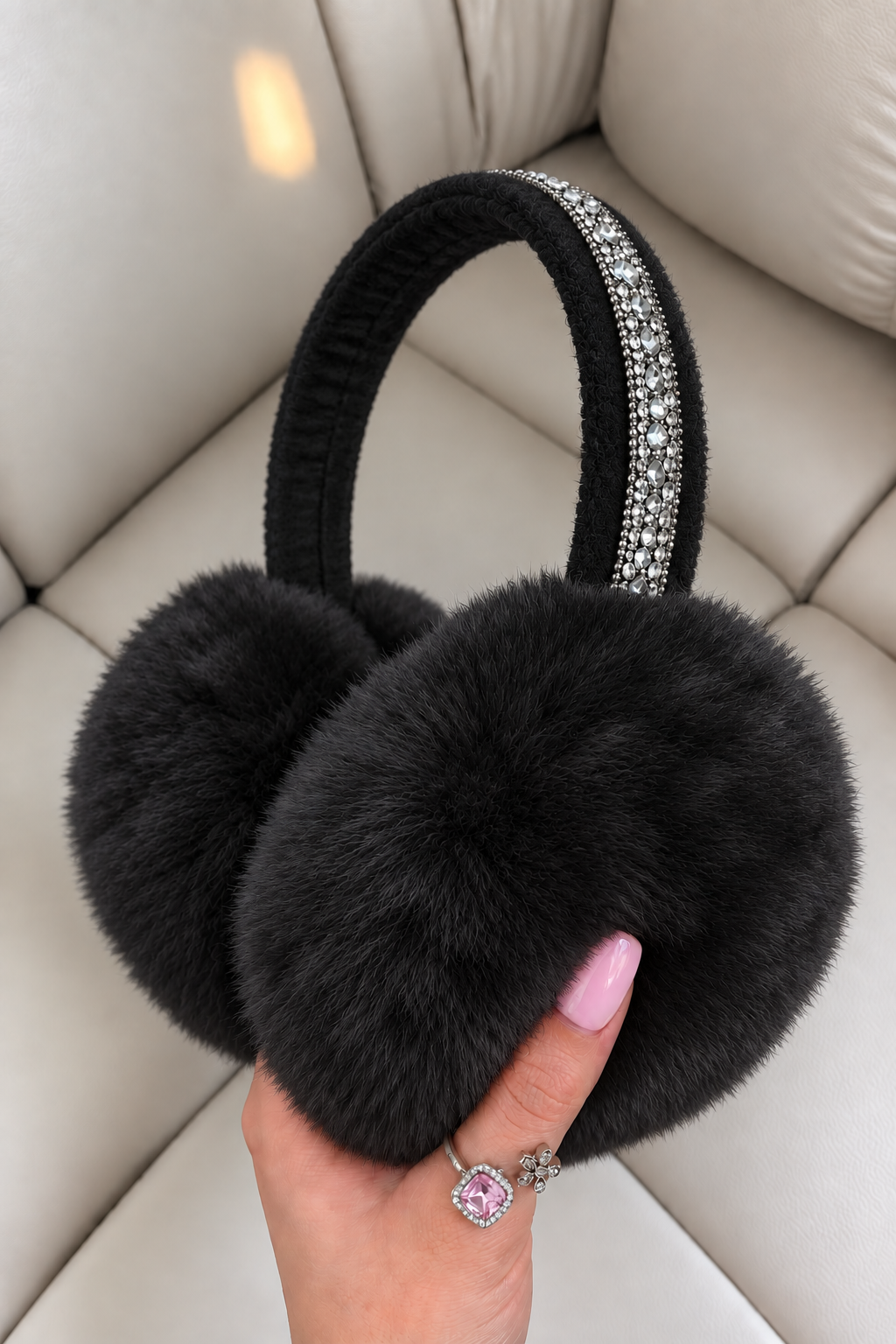 Black faux fur earmuffs with a sparkling rhinestone headband, held by a hand with pink manicured nails and a ring, sitting on a beige leather couch.