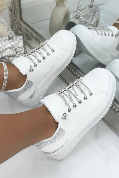 White sneakers with decorative laces and soles worn by a person, with a blurred background.