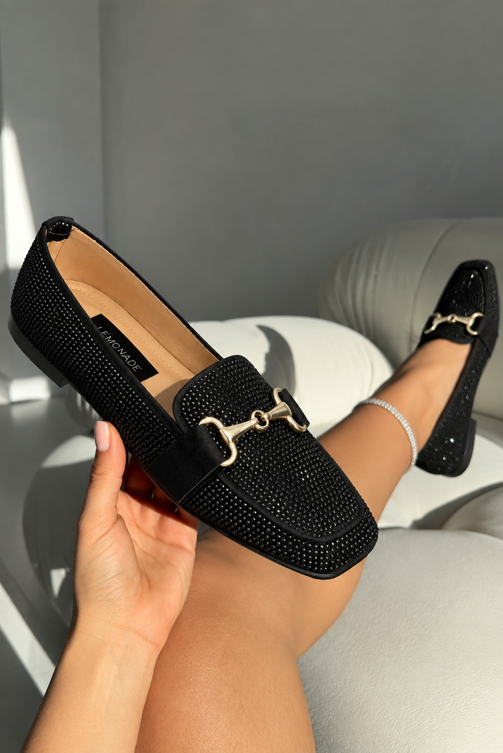 A person holds a black embellished loafer with a gold-tone horsebit detail, while wearing the matching shoe on their foot in a modern indoor setting.