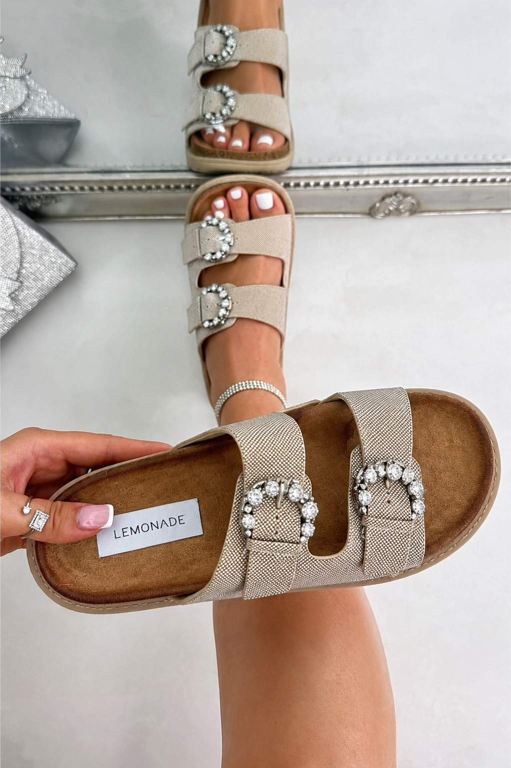 A person holds a pair of beige double-strap slide sandals with large rhinestone-embellished buckle details. The brand name &