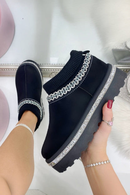 Black slip-on shoes with decorative straps held by a hand against a soft focus background
