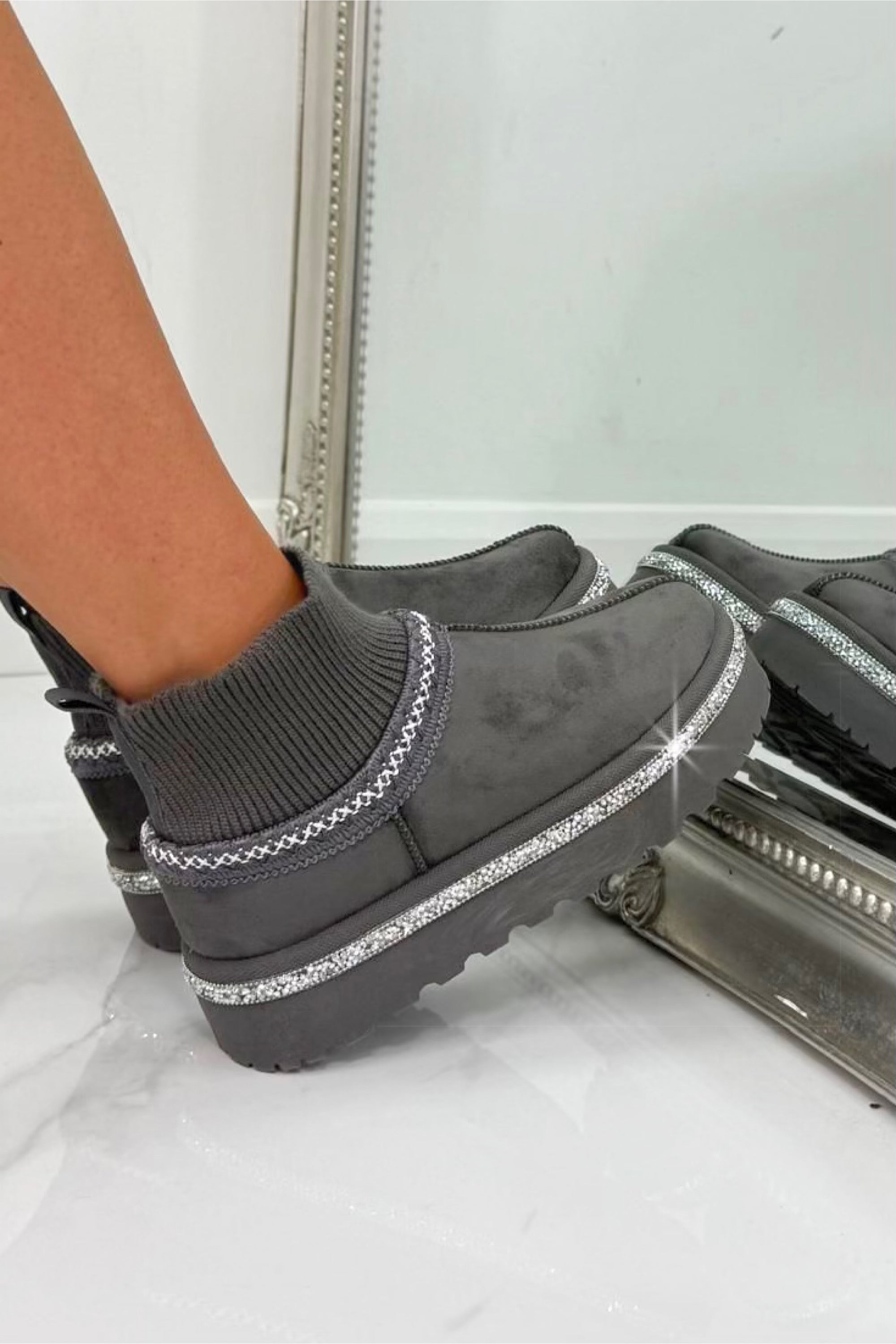 Gray slip-on boots with glittery trim worn by a person, standing on a white floor.