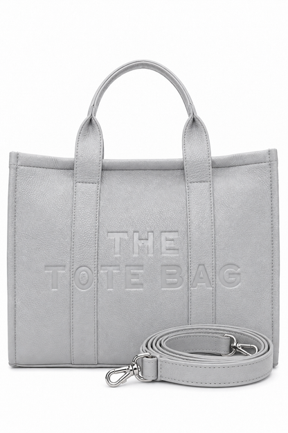 Large Grey faux leather structured tote bag with two rounded top handles, embossed. "THE TOTE BAG" lettering on the front, vertical stitched panels. Pictured on a white background.  With grey-tone strap pictured in front of the bag.