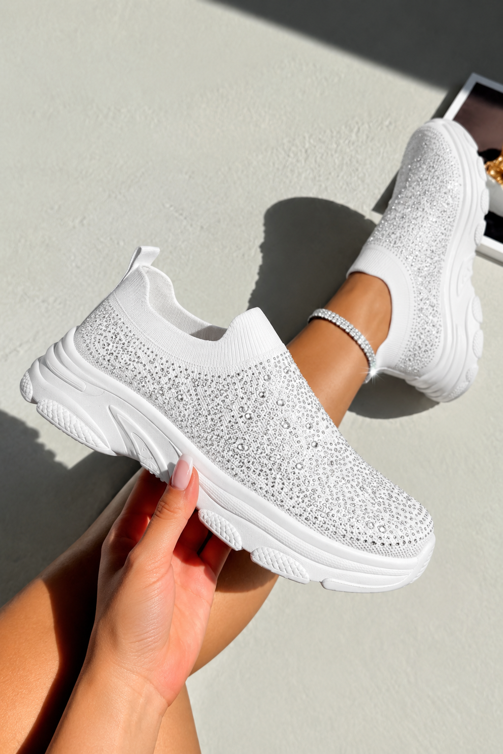 A pair of chunky white slip-on sneakers covered in sparkling rhinestones, shown being worn with one foot raised and the other held by a hand against a light gray wall.