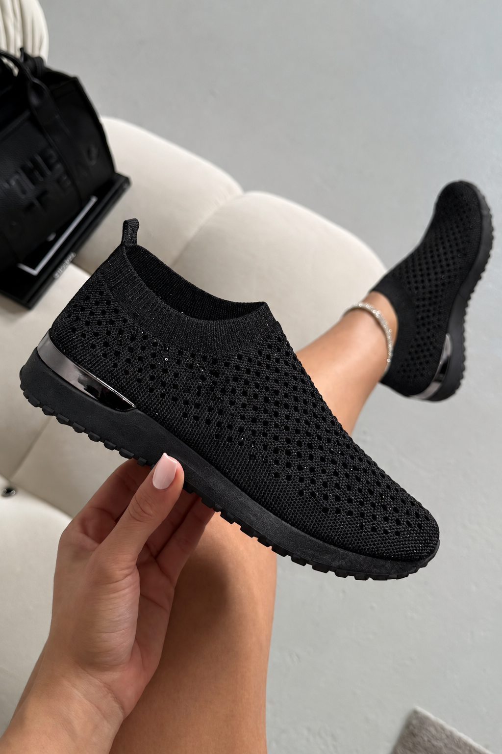 A person holds up a black knit slip-on sneaker while wearing the same shoe, with one foot raised. The shoe has a breathable perforated upper and a black sole, shown in a casual indoor setting with a light-colored chair and a black bag in the background.