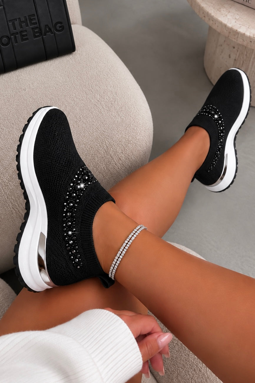 A person wearing black knit slip-on sneakers with silver rhinestone embellishments and chunky white soles, seated on a beige upholstered chair.