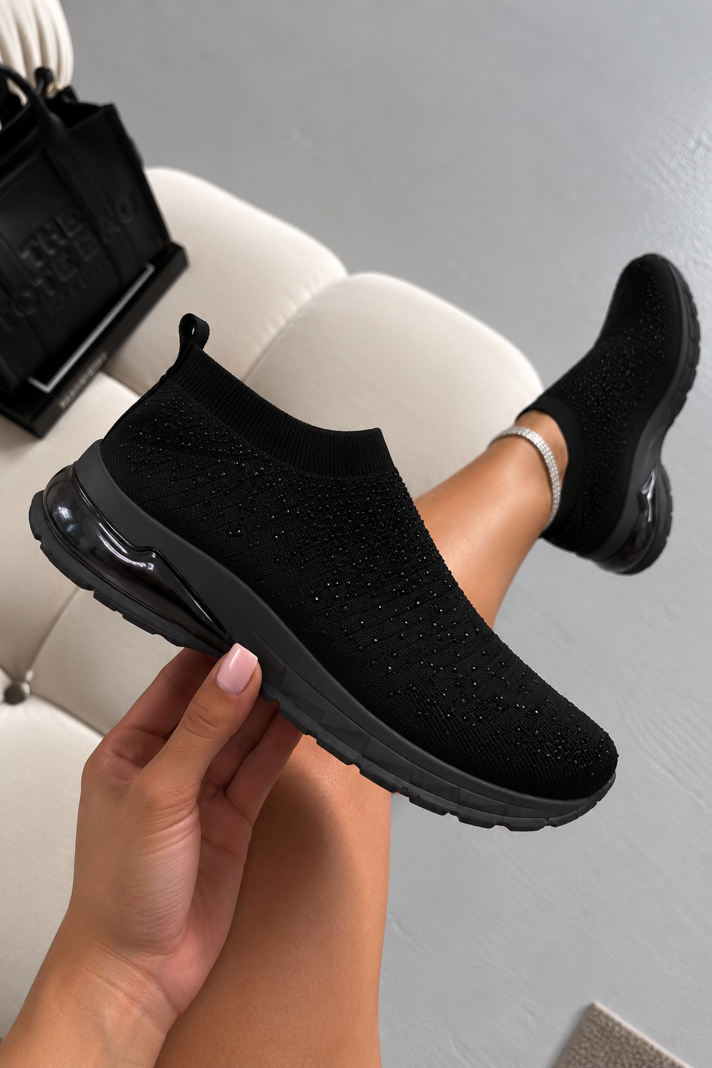 A person holds up a black slip-on knit sneaker with a chunky black sole, while another matching shoe is worn on their foot, set against a light gray background with a white chair.