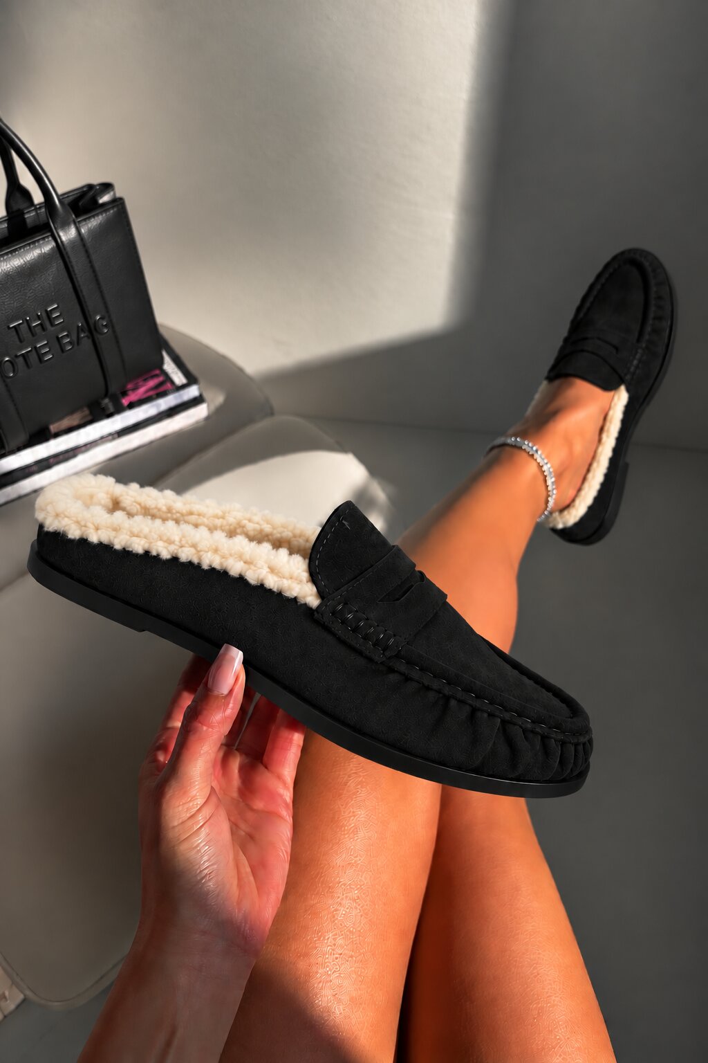 A person wearing black suede slip-on loafers with cream sherpa lining, shown with one shoe being held by a hand and the other resting on their foot. A black handbag is partially visible in the background on a light-colored chair.