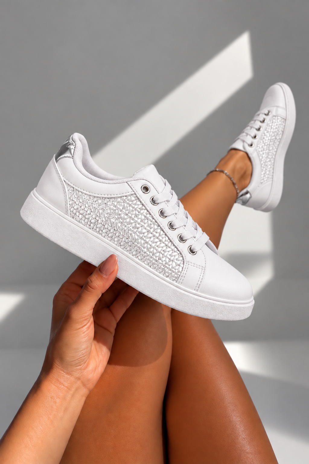 A pair of white low-top sneakers with textured silver mesh panels on the sides, held by a hand with crossed legs visible in the background against a gray wall with geometric shadows.