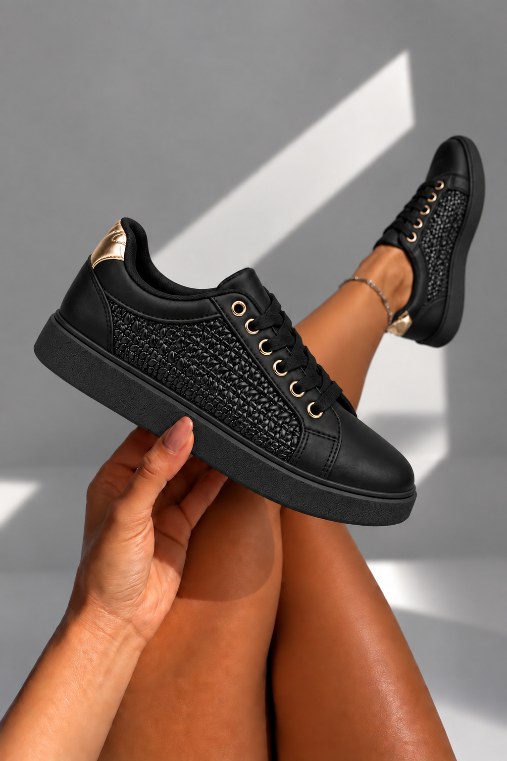 A pair of black low-top sneakers with textured knit side panels, black laces, gold-tone eyelets, and metallic gold heel accents, shown on a person&