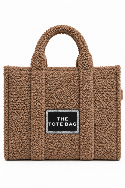 A brown teddy fleece tote bag with dual top handles and a black rectangular label on the front that reads &