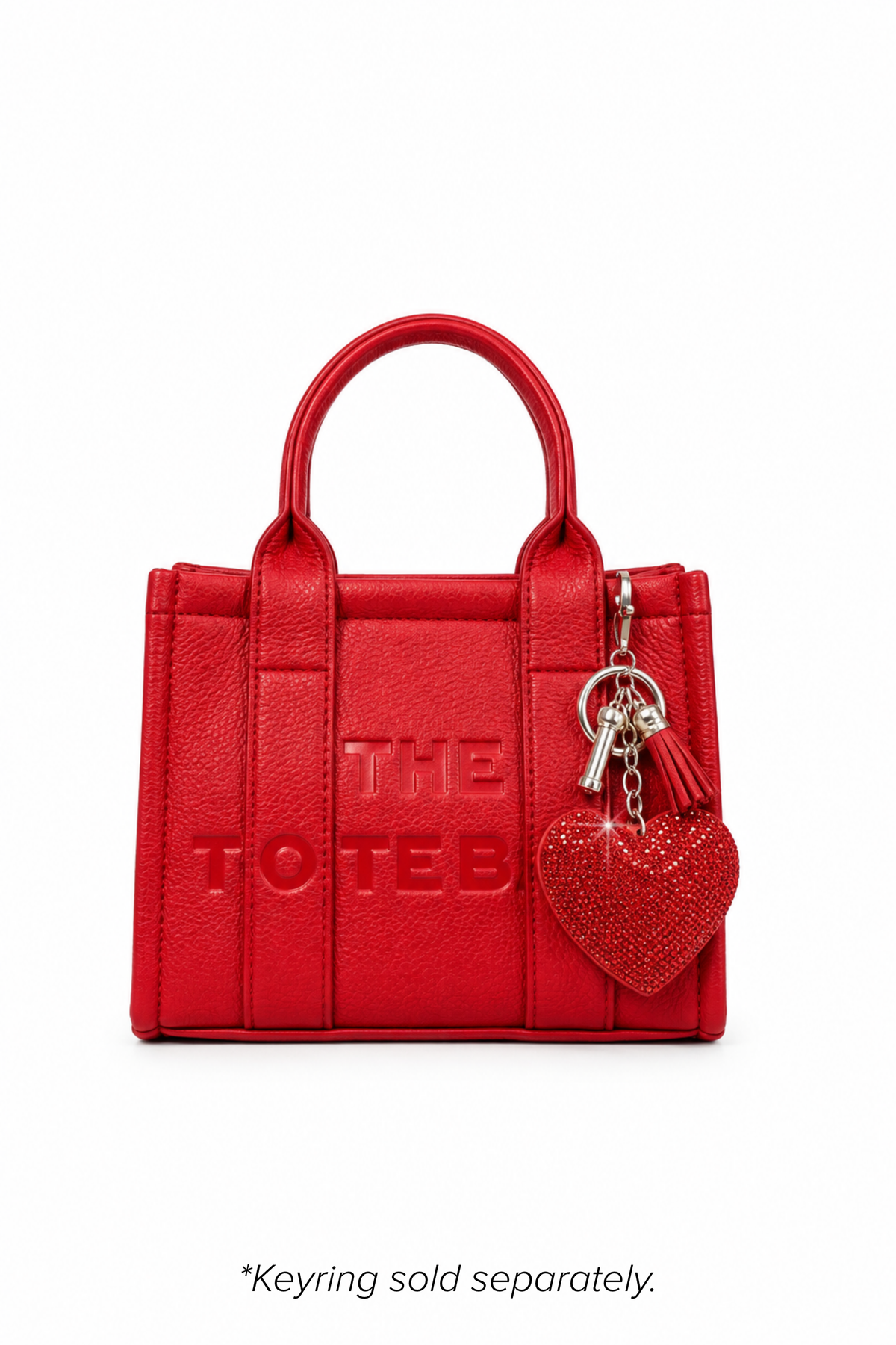 Red faux-leather handbag with rounded top handles, structured rectangular shape vertical stitched panels and embossed "THE TOTE BAG" lettering on the front. Features a top zip fastening and a silver tone hardware. A red glitter heart key-ring is attached to the side. Key-ring sold separately.