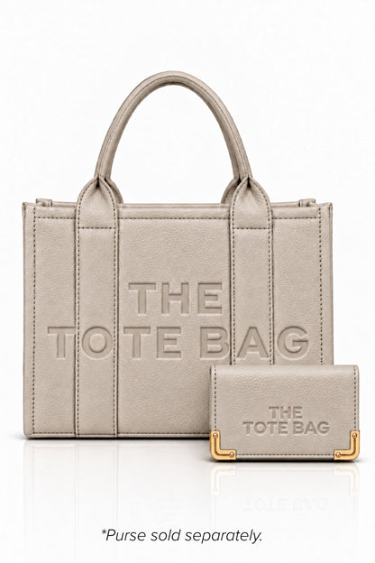 Beige faux leather tote bag with two rounded top handles, embossed "THE TOTE BAG" lettering on the front, contrast stitching. gold tone hardware and a detachable adjustable black shoulder strap. With a beige TOTE purse pictured in front of bag.