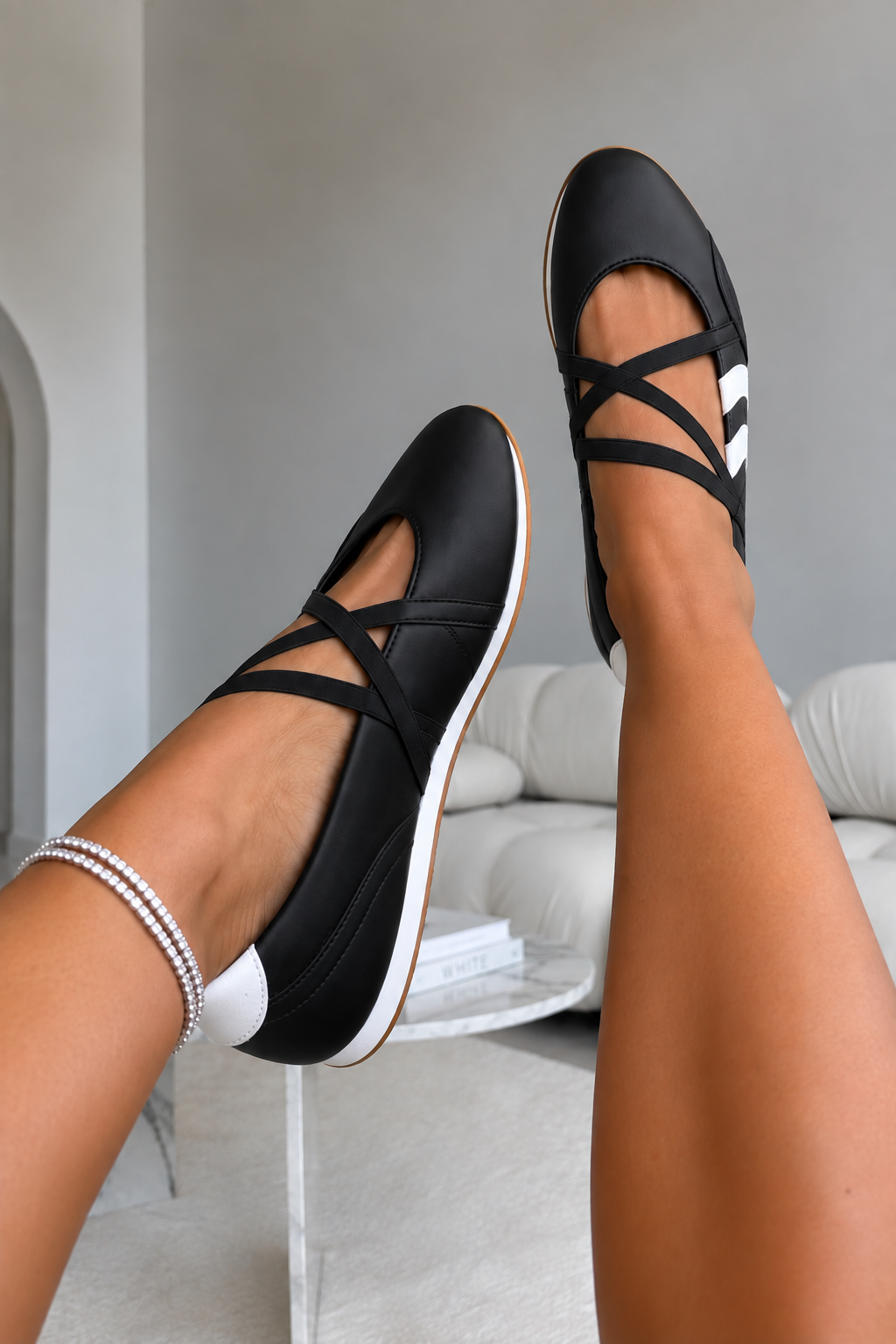 A person wearing black strappy ballet flats with white side accents and white soles, shown with both legs raised in a modern indoor setting with a light gray sofa in the background.