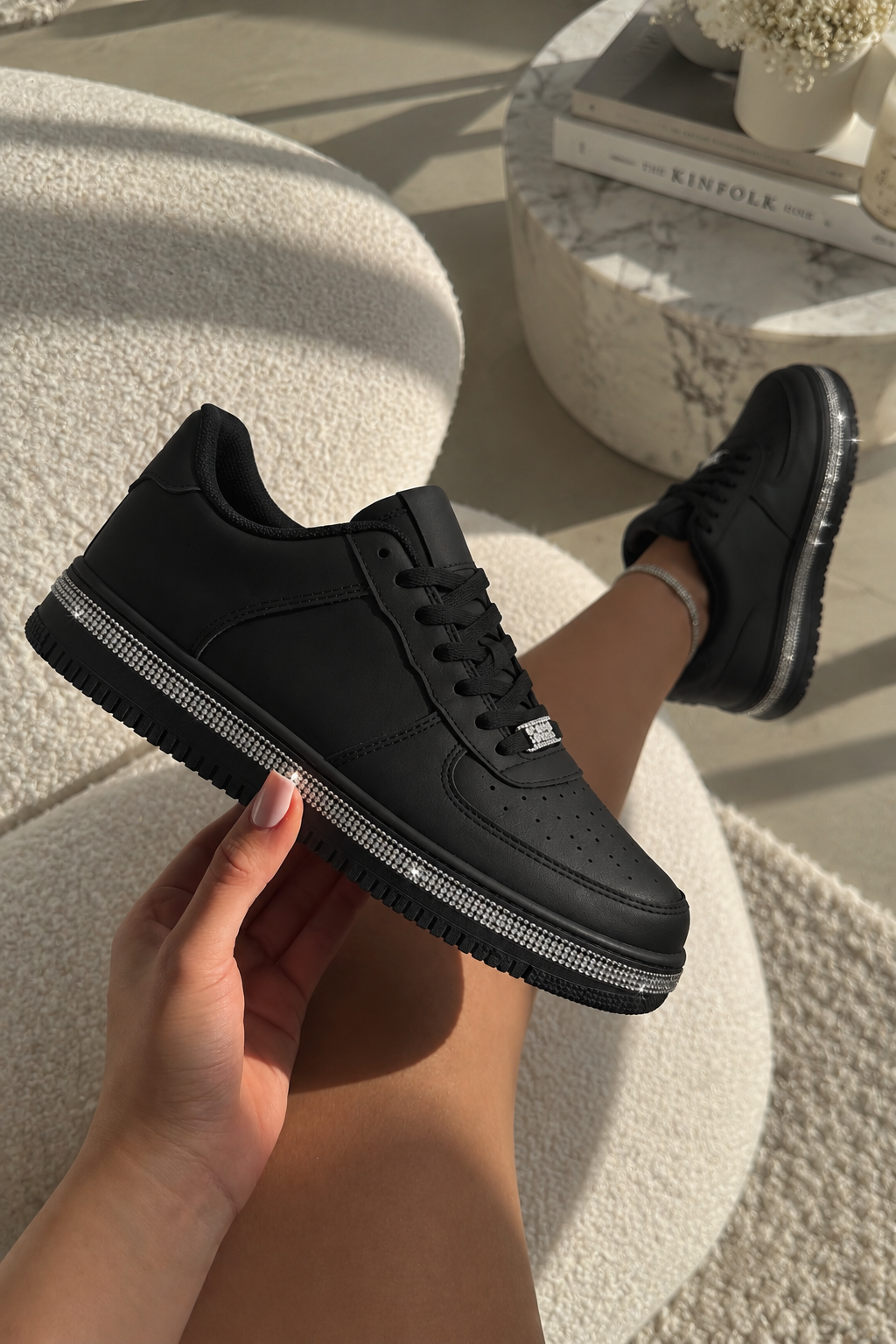 A person holds a black low-top sneaker with a silver rhinestone-embellished sole edge in a bright indoor setting, with another matching shoe visible on their foot.