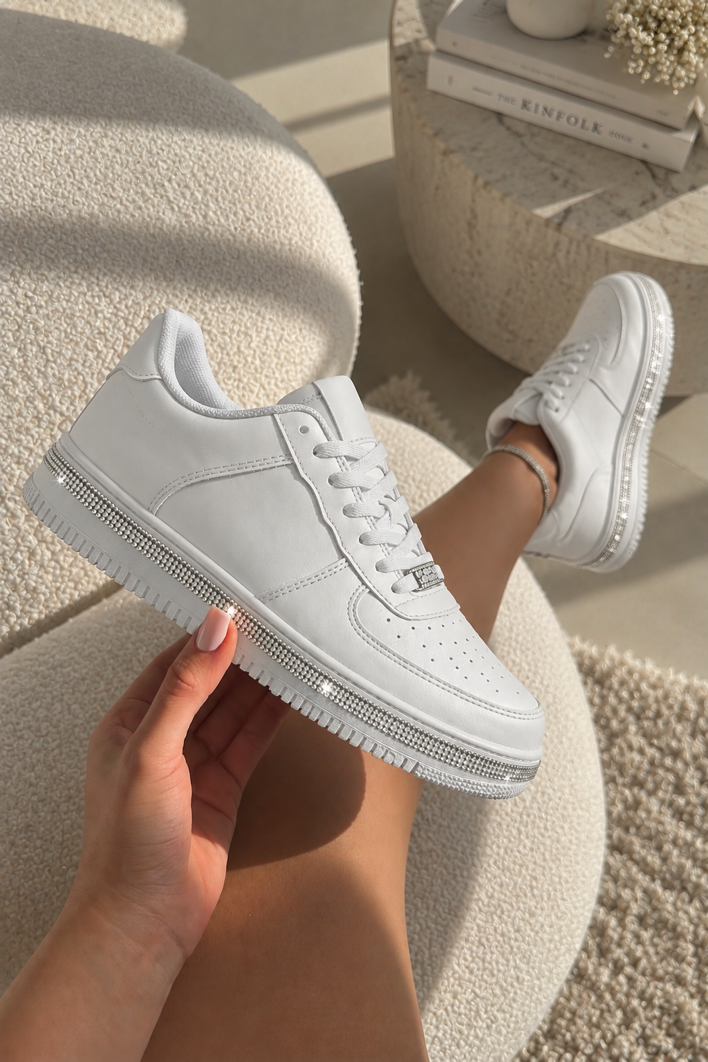A pair of white low-top sneakers with rhinestone-embellished soles, shown being held by a hand and worn on a foot in a bright, sunlit indoor setting.