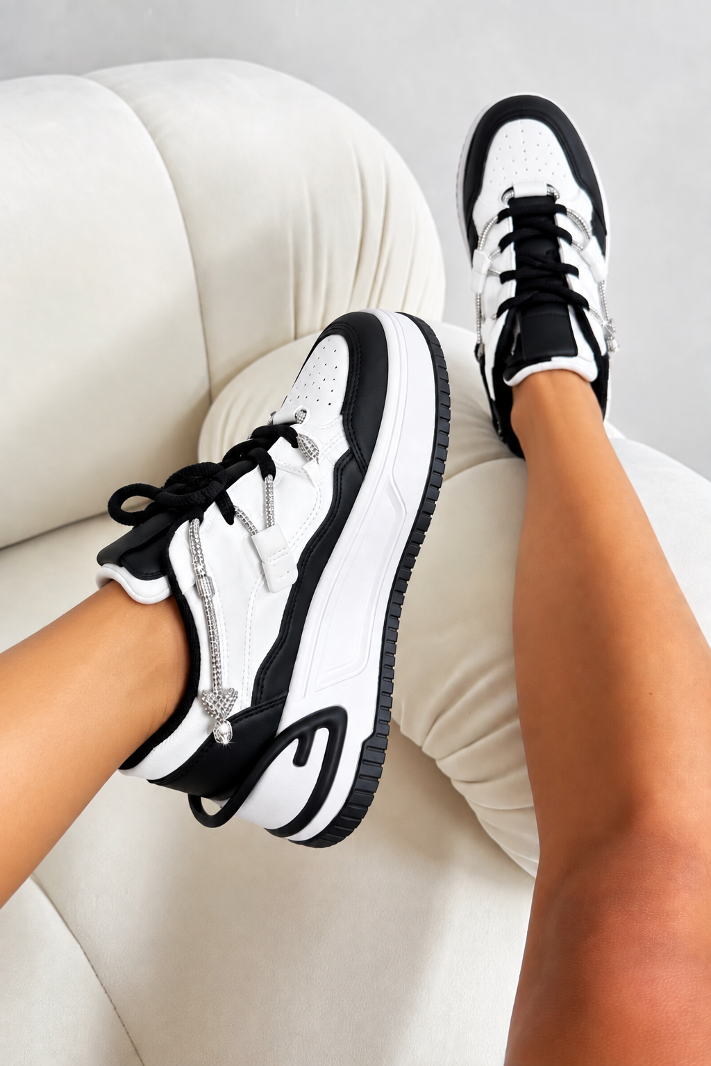 A pair of black and white platform sneakers with silver rhinestone zipper details, shown being worn on feet resting on a cream-colored sofa.