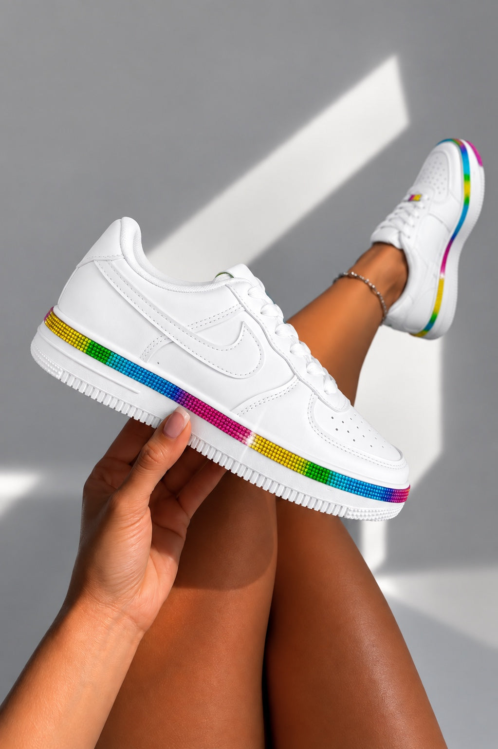 A white low-top sneaker with a rainbow-colored stripe along the midsole and outsole, held by a hand against a gray background with diagonal shadows. The model&