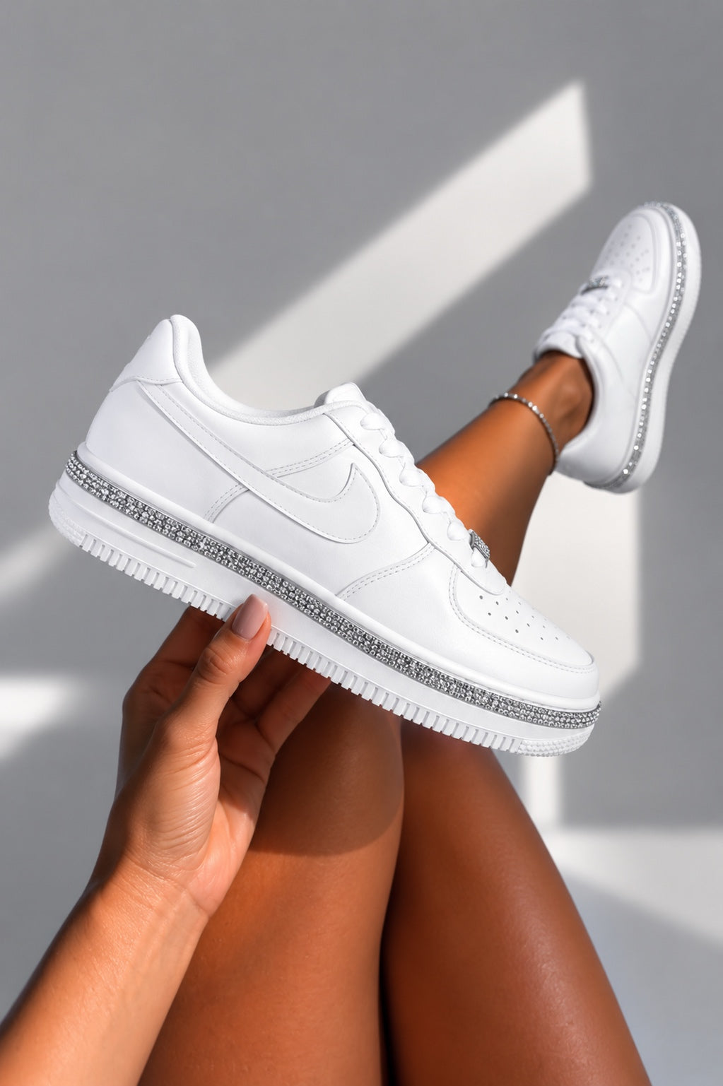 A white sneaker with silver rhinestone trim around the midsole, held by a hand with the other leg bent up wearing the same shoe. The background is a clean gray wall with diagonal shadows.