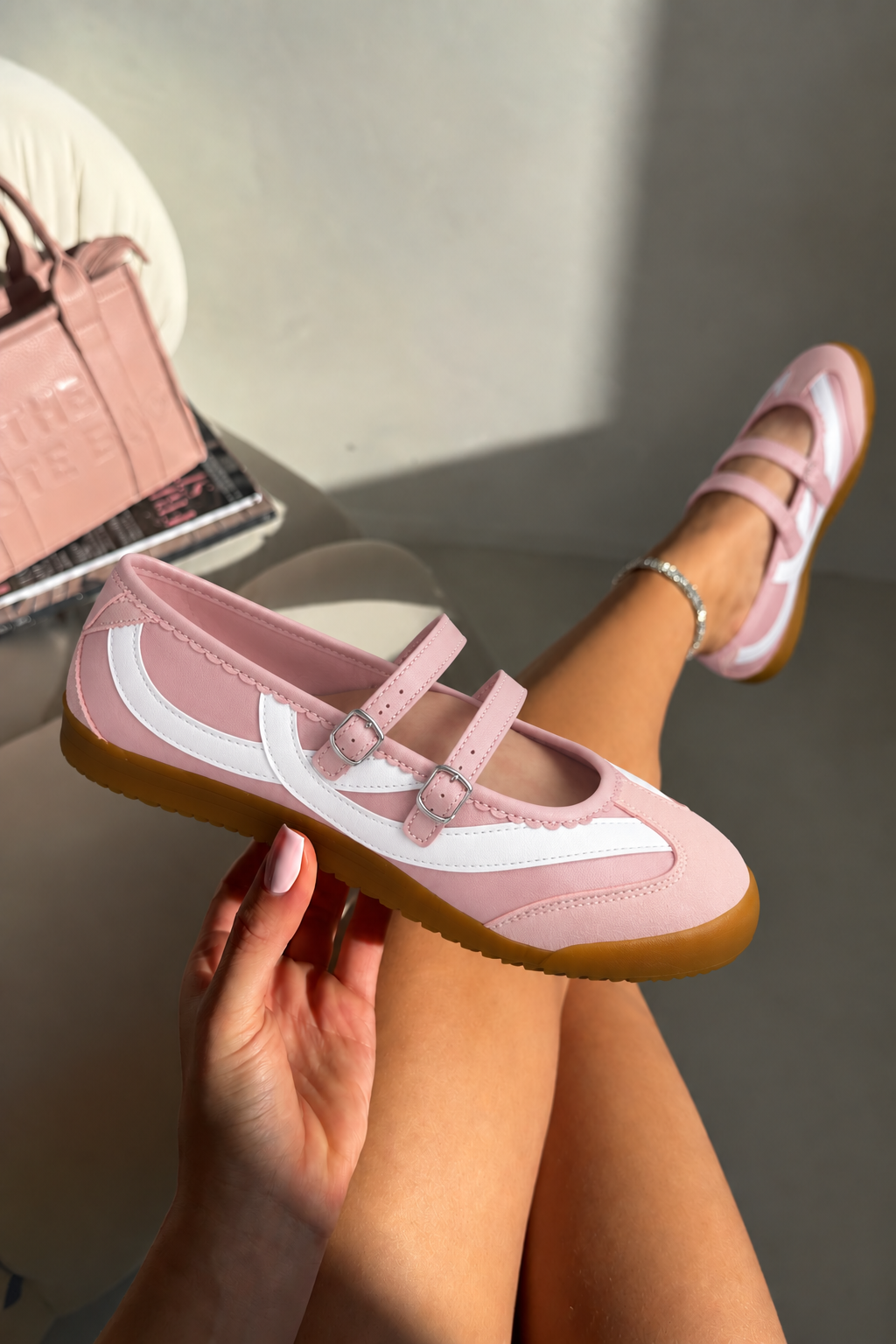 A person holds a light pink and white Mary Jane shoe in one hand while wearing the same shoe with their legs extended, in a sunlit indoor setting with a chair and handbag in the background.
